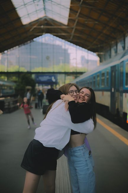 This image features two young women embracing and smiling joyfully at Walthamstow Central station. One woman has light brown hair, wears glasses, a white jacket, and black shorts, while the other has 