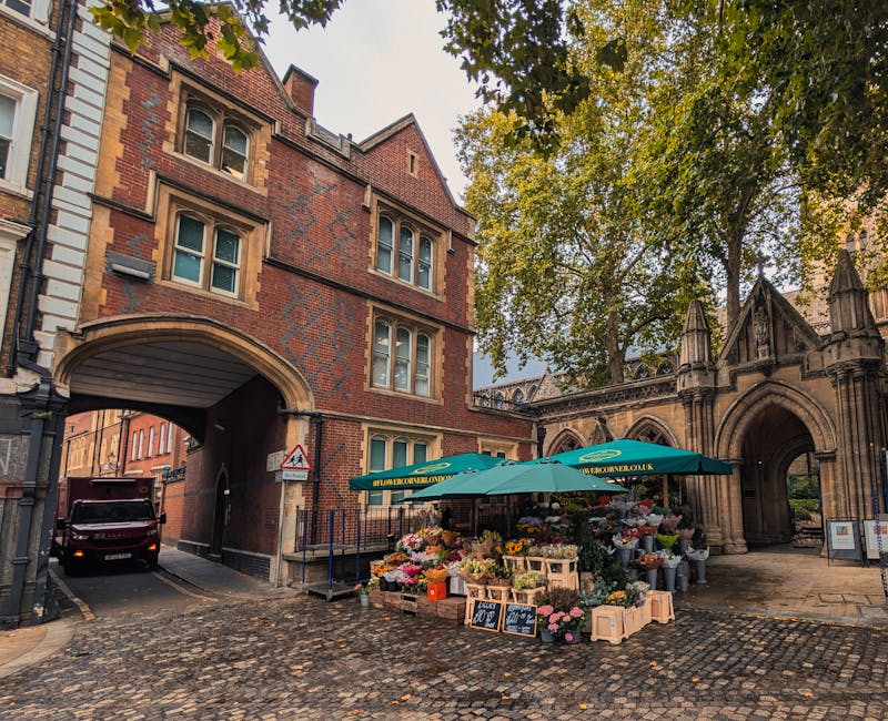 A street scene in Walthamstow featuring a flower stall positioned outside a historical brick building with arched gateways. The stall is shaded by a large green umbrella branded with the URL 'walthams