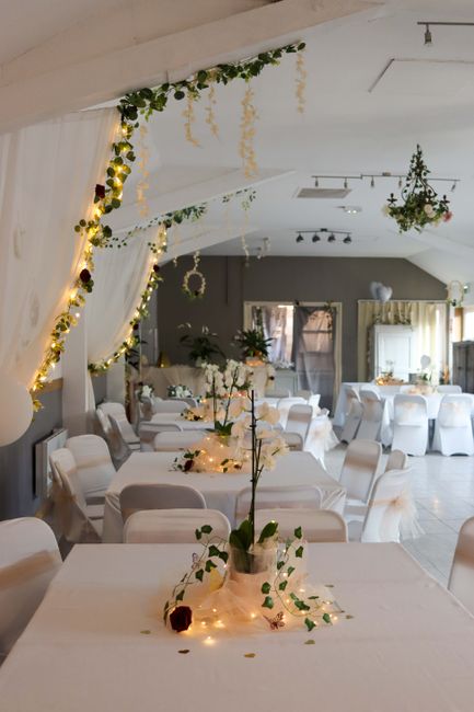 Interior view of a wedding reception venue decorated with floral arrangements and string lights. White tablecloths cover multiple round tables adorned with delicate floral centrepieces featuring white
